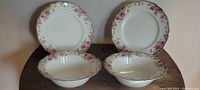 Four plates shown: two open vegetable dishes (deep bowls) and two dinner plates. All have off-white background with rose and foliage decoration on the rims, gold gilding evident.