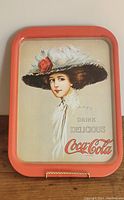 Front view of the Coca Cola collectible metal serving tray showing the vintage woman illustration with text "Drink Delicious Coca-Cola." Tray is red-orange edged with a beige background.