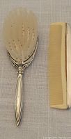 Full view of baby hairbrush with ornate sterling silver handle and soft bristles next to baby comb with plastic teeth and silver backing.
