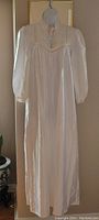 Full front view of the vintage white satin nightgown hanging on a dress form against a plain background, showing its full length and shape with long sleeves and subtle sheen.