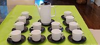 12 white cups and 12 black saucers arranged on table with coffee pot at center, showing full set layout
