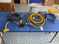 Wide view of all five electrical cords laid on a blue table, showing composite of different types and colors, primarily black and yellow with 30 amp plugs and connectors.