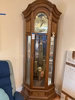 Full frontal view of the complete grandfather clock showing wooden arched top, glass front door, and brass pendulum inside.