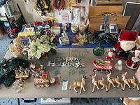 Overall view of assortment of Christmas decorations on table including fiber optic angel, lights, tabletop decor, Santas, reindeer, wreaths, garland, and Happy Holidays sign.