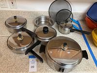 Photo of assorted stainless steel pots and pans with lids along with a small stainless steel dish and two splatter screens on a countertop.
