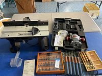 Overall view of router table, router kit in case, assortment of wood chisels, and router bits case on a table