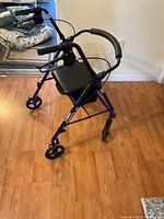 Blue rollator walker with padded seat, backrest, four wheels, and hand brakes, set up in room with wooden floor.