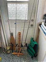 Photo showing four fishing rods with reels standing up next to a green life jacket and a wooden paddle, against a white door and a blue painted floor.