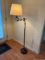 Full view of the black iron floor lamp standing on a wooden floor beside a wall and doorway. Shows lamp lit with fabric shade and visible power cable.