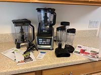 Full view of Ninja Auto-IQ blender base with two blender jars, lids, tamper, other accessories, and manuals on kitchen counter.