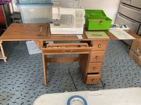 Wide view showing the sewing machine set in the wooden cabinet with side panels extended, drawers, owner's manuals, and green sewing basket on cabinet surface.