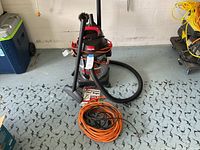 Shop-Vac vacuum with attached hose, black wand nozzle, and orange extension cord coiled in front with wall and garage floor background.