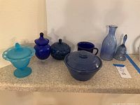 Image showing a variety of vintage blue glassware and stoneware items arranged on countertop, including covered dishes, carafe, bell and cup.