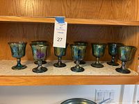 Nine iridescent carnival glass goblets with grape design arranged in a row on a shelf showing color reflections and structural details.