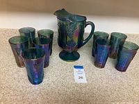 Full set of pitcher and nine glasses on countertop showing iridescent surface and grape motif.