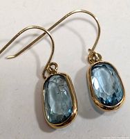Pair of delicate 14K gold earrings featuring oval faceted blue topaz gemstones in bezel settings, shown flat on white surface.
