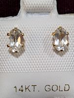 Front view of a pair of oval white topaz stones set in 14K yellow gold prong settings on an earring display card marked '14KT. GOLD'.
