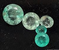 Close-up photo of five round cut emerald stones in green hues with visible inclusions, placed on dark fabric background.