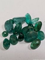 Multiple polished green emerald gemstones in mixed shapes and sizes arranged on a plain white background.