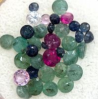 Close up of assorted green emerald, red ruby, blue sapphire and other small faceted and cabochon cut gemstones on a white background.