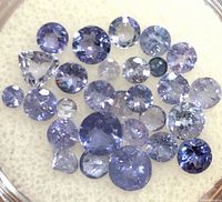 Close-up of around 30 loose tanzanite gemstones in round and pear cuts, various sizes, violet-blue color.