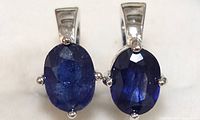 Close-up image showing both silver earrings with oval faceted blue sapphire stones set with four silver prongs each.