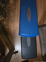 Photo shows the top view of two fitness steppers stacked on top of each other; the blue one on top and the black one underneath, both featuring a diamond patterned nonslip surface and silver oval panels in the center.