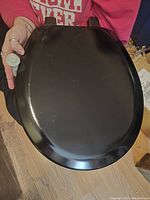 View of the black toilet seat showing top surface and part of the mounting hardware held by a person