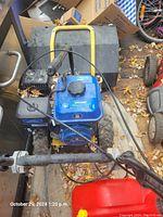 Photo showing the gas-powered sweeper from the rear, highlighting the blue engine, black control cables, and thick rubber tires with a rugged pattern. Leaves and debris are visible around the machine.
