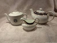 Three items side by side showing white porcelain with gold and blue pattern: a teapot, a matching creamer, and another pot with a lid and handles.
