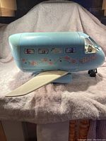Side profile of vintage Barbie airplane toy. Light blue plastic body with floral and butterfly decals. Several window openings. Plane wing extended. Toy stands on landing gear wheels.