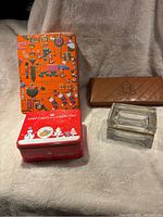 Full view of 2 tins, 1 glass box, and 1 brown leather case on a beige fabric background.