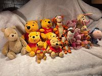 Full view of the entire Winnie the Pooh themed stuffed animal collection including Pooh in various styles, Tigger, Piglet, Rabbit, Eeyore, and Roo, all arranged on a beige blanket.