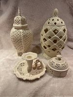 Four creamware ceramic decorative items displayed on a soft textured background: tall urn with latticework lid, small lidded bowl, chamberstick candle holder, hexagonal container with removable inner liner.