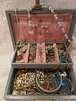 Full view of open gray box with pink lining showing a collection of various jewelry pieces including rings, necklaces, bracelets, brooches, and a watch in compartments and loosely packed below.