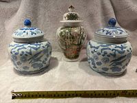 Front view of all three ceramic lidded ginger pots, two white and blue and one decorated with floral and garden landscape scene.