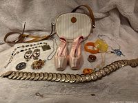 Photo of all items displayed together on beige fabric showing ballet shoes, handbag, gold-tone belt, various bracelets, and jewelry pieces.