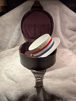 Vintage white straw hat with flat top and tricolor ribbon inside open brown leather hat box on a white cloth background.