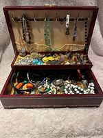 Open red jewelry box showing two levels filled with assorted jewelry items including necklaces, rings, earrings, bracelets, brooches and pins.