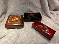 Photo showing all three musical jewelry boxes together on a soft beige backdrop, highlighting wooden and lacquered materials and floral designs.