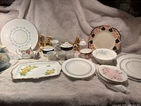 Full lot showing assorted china including plates, bowls, cups, two gold candle holders on a light cloth background