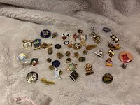 Photo showing an assortment of various lapel pins on a beige blanket including gold-toned pins, colorful pins, and emblem pins.