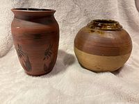 Two pottery vases placed side by side on a soft surface, showing one reddish-brown vase with floral motifs beside a round two-tone brown vase.