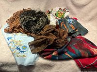 Lot of six silk scarves with varied prints and colors arranged on a beige fabric background.