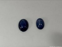 Two synthetic star sapphires placed on a flat surface showing their star asterism effect in oval cabochon form.