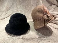 Two vintage women's hats on cream plush fabric: black faux fur cloche and beige wool hat with bow and feather detail.