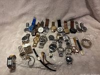 Overview of assortment of 28 analog wristwatches with various strap materials and colors arranged on fabric surface