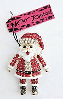 Santa Claus shaped pendant or brooch decorated with red and clear rhinestones, showing detail and attached Betsey Johnson tag.