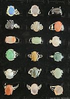Photo of 18 fashion rings arranged in rows of 3 across and 6 down on a black velvet display, showing various colors and styles of oval stones in silver-tone settings.