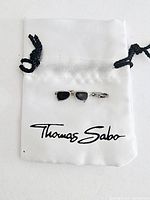 Charm shaped like sunglasses with black enamel lenses resting on a white fabric gift bag with 'Thomas Sabo' logo.